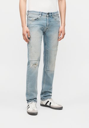 Personne portant un jean délavé bleu clair avec un patch et des détails usés, associé à des baskets blanches avec des bandes noires.