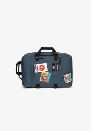 Blue fabric duffle bag with black straps, featuring multiple colorful patches and logos on the front. Zipper closure at the top.
