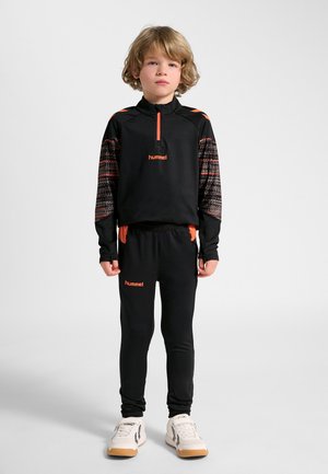 Garçon aux cheveux châtain clair debout, portant un survêtement de sport noir et orange et des baskets blanches sur un fond clair uni.