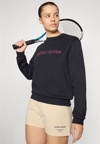 Sweatshirt preto com "BJÖRN BORG" em bordado lilás, combinado com calções bege com estampado de logo. Inclui uma raquete de ténis como acessório.