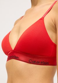 Raudonas medvilninis braletė su giliu V formos iškirpimu, plonomis petnešėlėmis ir kontrastingu Calvin Klein logotipu ant elastingos juostos. Glotni tekstūra.