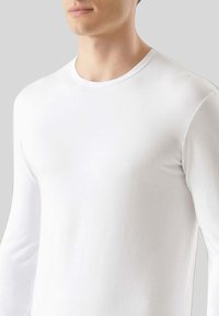 Camicia bianca a maniche lunghe realizzata in tessuto morbido. Caratterizzata da un collo a giro e una consistenza liscia. Design aderente senza motivi o dettagli.