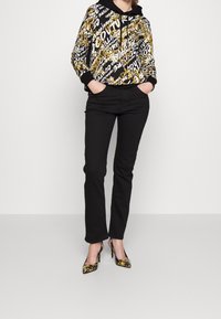 Mikina s černým a zlatým vzorem a stahovací kapucí, spárovaná s úzkými černými kalhotami a černými vysokými podpatky se zlatým květinovým designem.