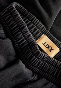 Schwarzer elastischer Taillenbund mit geraffter Textur, ausgestattet mit einem beige Stofflabel, auf dem "JJXX" in Schwarz gedruckt ist. Weiches Material mit glatter Oberfläche.