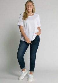 T-shirt bianca a maniche corte, vestibilità rilassata, abbinata a jeans blu scuro. Il modello indossa scarpe da ginnastica bianche e una collana di perline. Sfondo neutro.