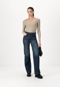 Beige Langarm-Bodysuit kombiniert mit hochgeschnittenen, dunkelblauen, weit geschnittenen Jeans. Modell hält eine schwarze Tasche und steht vor einem weißen Hintergrund.
