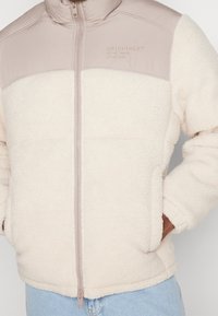 Fleecejacka med en beige övre del och en krämfärgad nedre del, med full dragkedja, hög krage och diskret logotyp på bröstet.
