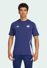 Navyblå kortærmet t-shirt med pink detaljer, prydet med et hane-logo og tre striber på skuldrene. Fremstillet af åndbart stof.