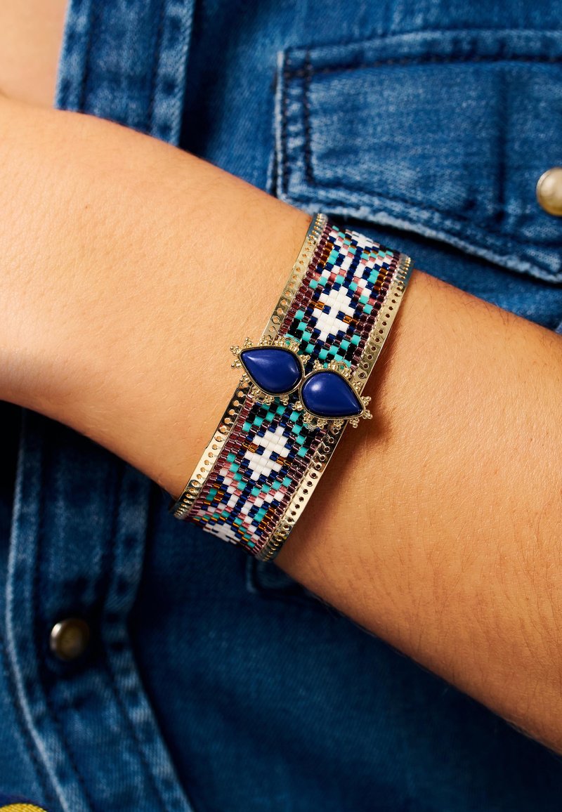 Bracelet en perles avec une base en métal doré, présentant des motifs géométriques multicolores et deux pierres en forme de goutte d'eau bleues en accent.