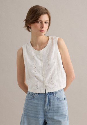 Mujer joven con cabello corto y castaño que lleva una blusa blanca sin mangas con botones, con recortes florales y vaqueros de talle alto en azul claro.
