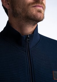 Navy zip-up jacket featuring ribbed texture, high collar, and subtle logo patch. Zipper is centrally placed on the front.