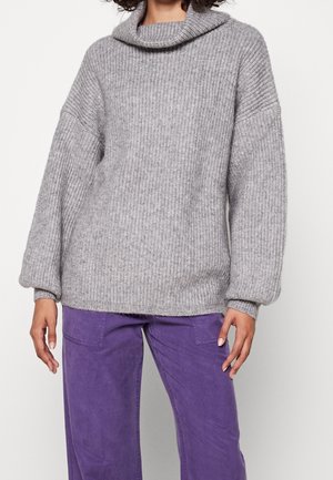 Personne portant un pull gris ample en maille côtelée avec un col bénitier et des manches longues, associé à un pantalon violet.