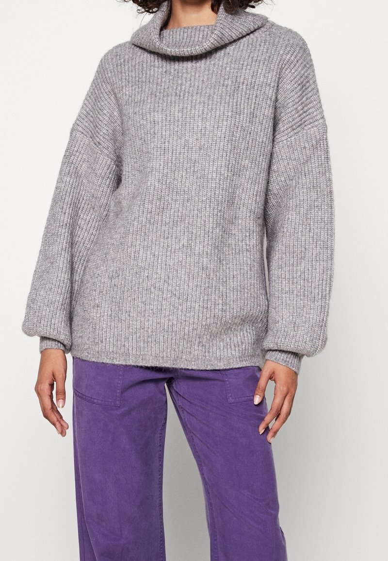 Personne portant un pull gris ample en maille côtelée avec un col bénitier et des manches longues, associé à un pantalon violet.