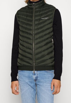 Mężczyzna noszący ciemnozieloną pikowaną kamizelkę na zamek, czarną koszulkę z długim rękawem i jasnoniebieskie dżinsy, logo Armani Exchange na piersi.
