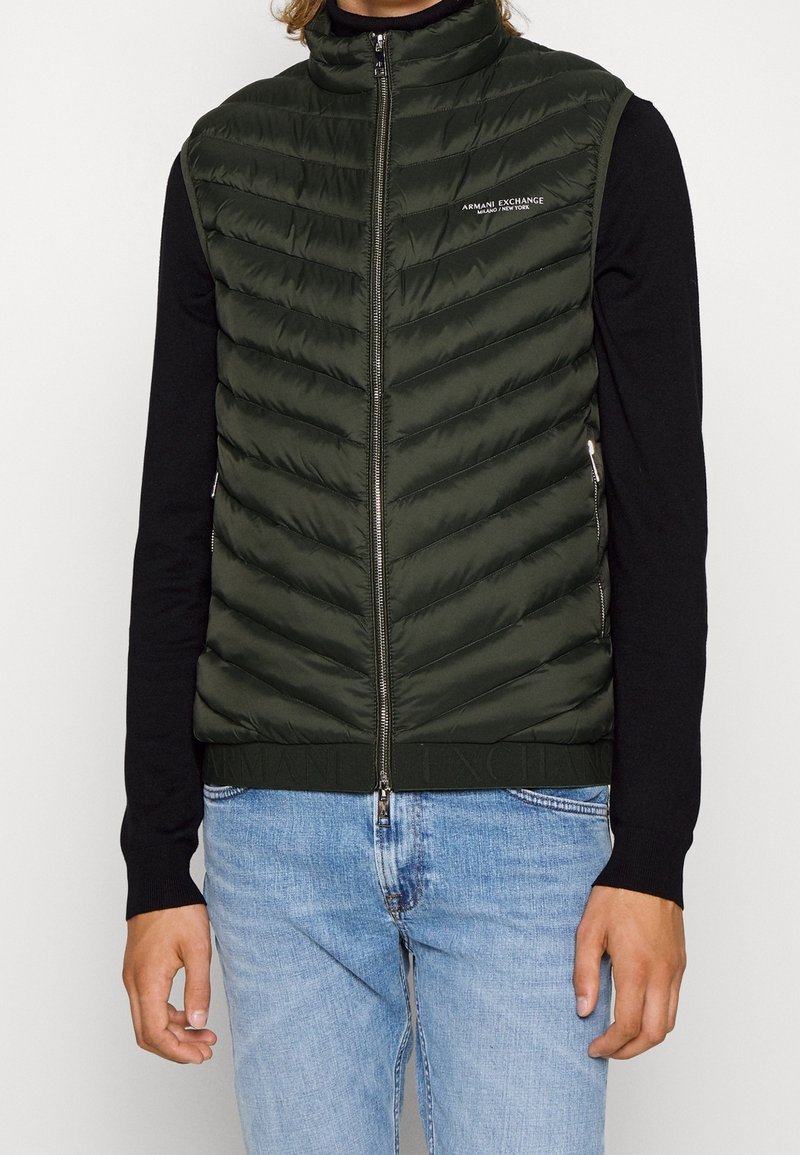 Homme portant une veste matelassée vert foncé zippée sur une chemise noire à manches longues et un jean bleu clair, logo Armani Exchange sur la poitrine.