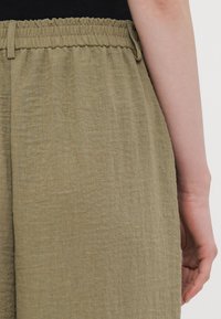 Pantalone od teksturirane tkanine u maslinasto zelenoj boji s nabranim elastičnim pojasom i koso postavljenim bočnim džepovima, s opuštenim krojem i laganom težinom.
