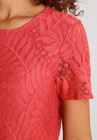 Koronkowa bluzka z krótkim rękawem w kolorze koralowym z wzorami kwiatowymi noszona na górnej części tułowia i ramieniu.