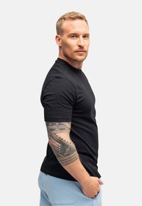 T-shirt nera a maniche corte in design aderente, indossata con pantaloni azzurri chiari, che mostra un tatuaggio dettagliato sul braccio del modello.