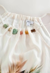 Haut blanc sans manches avec encolure froncée, orné de perles multicolores ressemblant à des pierres, avec une étiquette indiquant "NEXT Fabriqué en Inde".