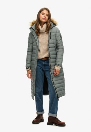 Manteau long rembourré en vert muted avec une capuche en fourrure, présentant une fermeture éclair, des coutures verticales et des poignets élastiques. Porté sur un pull en tricot beige et un jean bleu, associé à des bottes marron.