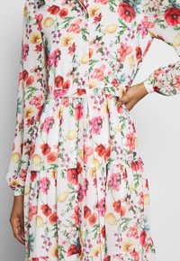 Blumenkleid mit weißem Hintergrund und mehrfarbigen Blumen, langen Ärmeln, tailliertem Mieder und einem gestuften Rockdesign.