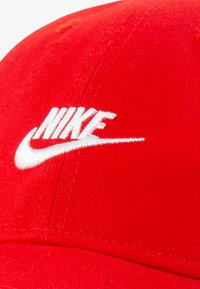 Sarkans auduma cepure ar izliekto malām. Priekšpusē ir balts izšūts "Nike" logotips un redzamas šuvju detaļas.