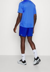 Hombre vestido con una camiseta deportiva azul y pantalones cortos, calcetines blancos y zapatillas de correr blancas y negras, de pie contra un fondo blanco.