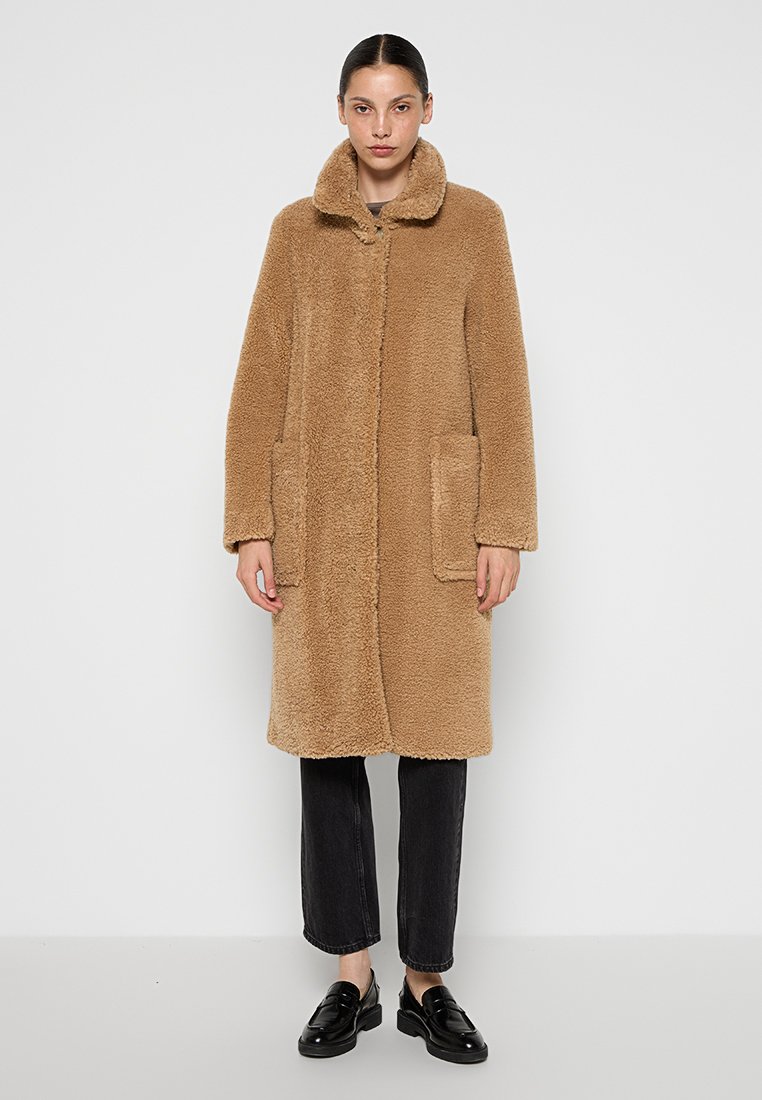 Un manteau en laine fluffy beige, avec un col, deux poches avant et une coupe droite. Porté sur un pantalon noir et des chaussures, sur un fond neutre.