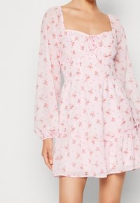 Vestido floral rosa con escote cuadrado, mangas largas y un dobladillo con volantes en capas. Confeccionado con una tela ligera y transparente con pequeñas flores delicadas.