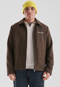 Mężczyzna w żółtej czapce beanie, brązowej kurtce zapinanej na zamek w wzór houndstooth, białym swetrze i ciemnych dżinsach, stojący na jednolitym tle.