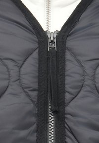 Melna pūšamā veste ar teksturētu lentu vidū, centrālu rāvējslēdzēju, izcelta ar kontrastējošu austu apmali un ar šūtu detaļu.