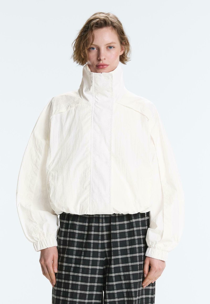 Femme aux cheveux blonds courts portant une veste blanche à col haut et un pantalon à carreaux noir et blanc, debout devant un fond uni.