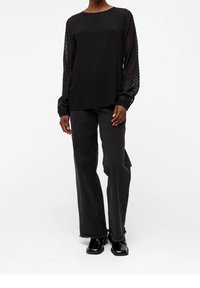 Top negro con mangas de malla texturizada, combinado con unos jeans anchos negros y zapatos brillantes, destacando un diseño en capas y una silueta moderna.