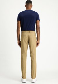 Pantaloni beige con vestibilità dritta, dotati di due tasche posteriori e una texture liscia. Indossati con una t-shirt blu navy e sneakers bianche.