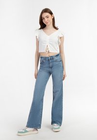 Hvid cropped top med flæser på ærmerne og en knude foran, sat sammen med lyseblå bell-bottom jeans og multikolore sneakers på en ensfarvet baggrund.