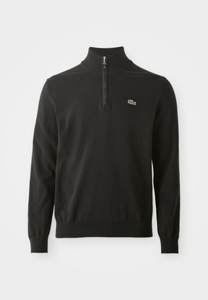 Svart genser med høy krage, med en halv glidelås og ribbestrikkede mansjetter. Inkluderer en liten grønn logo på brystet. Myk tekstur.