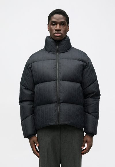 Doudoune noire avec un col haut, présentant un design matelassé et une fermeture éclair à l'avant. Le tissu a un motif subtil.