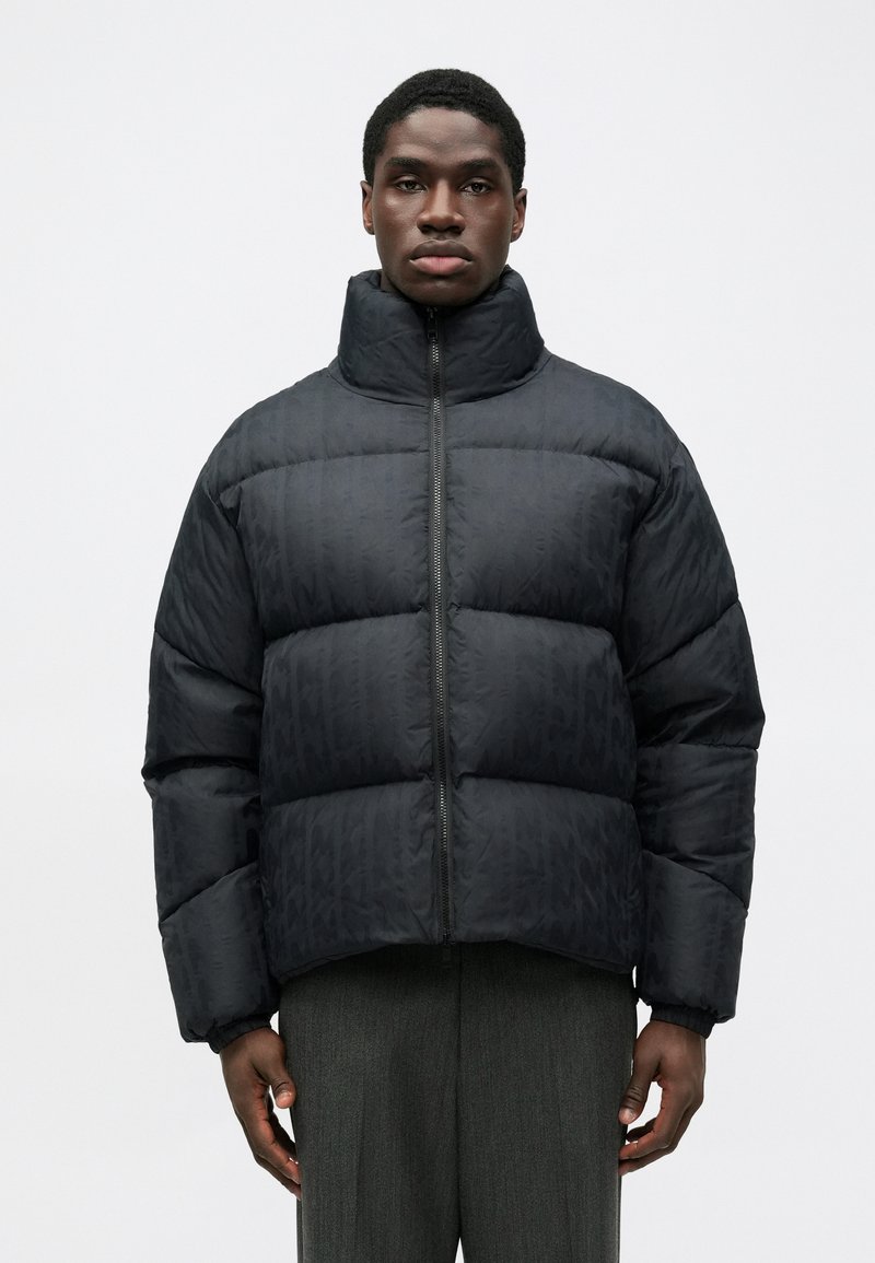 Jachetă puffer neagră cu guler înalt, având un design matlasat și închidere cu fermoar pe față. Materialul are un model subtil.