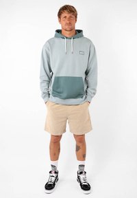 Sweatshirt à capuche bleu clair avec une poche vert foncé, associé à un short beige. Présente des cordons de serrage et un logo sur la poitrine. Baskets noires.