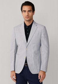 Muškarac u svijetlosivom blejzeru preko tamne polo majice i tamnoplavim hlačama stoji ispred jednobojne pozadine.