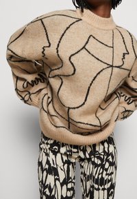 Beige Pullover mit schwarzem abstraktem Linienstil, gerippten Bündchen und Saum. Kombiniert mit gemusterten schwarzen und cremefarbenen Hosen, die kräftige Kritzeleien aufweisen.