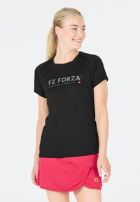 Leende kvinna med blont hästsvans som bär en svart FZ Forza-t-shirt och en röd sportkjol stående mot vit bakgrund.