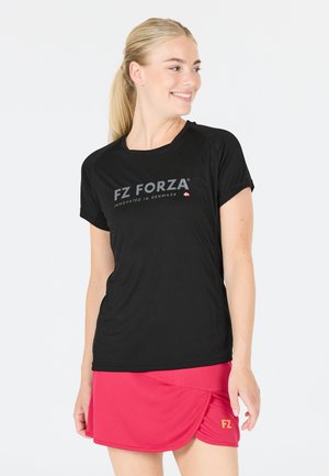 Smilende kvinde med blond hestehale iført en sort FZ Forza t-shirt og en rød sportsnederdel stående mod en hvid baggrund.