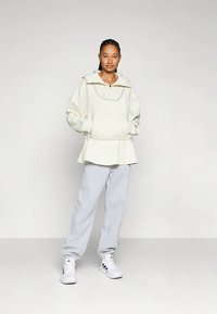 Γυναίκα στέκεται με τα χέρια στις τσέπες, φορώντας υπέρμετρο μακριά φούτερ σε ελεγχόμενο λευκό χρώμα, ανοιχτό γκρι παντελόνι φόρμας και λευκά αθλητικά παπούτσια Adidas, με φόντο απλή επιφάνεια.