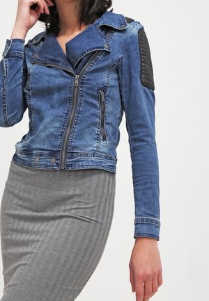 Kobieta w niebieskiej dżinsowej kurtce typu biker z czarnymi pikowanymi łatami na ramionach oraz dopasowaną szarą prążkowaną spódnicą na jednolitym tle.