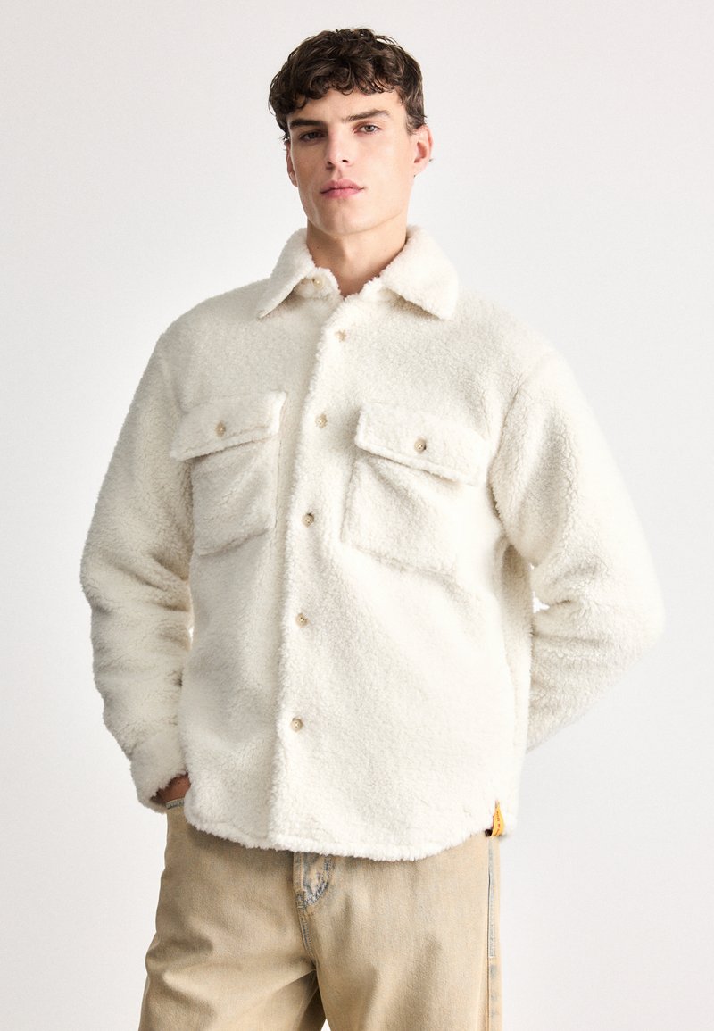 Hvit fleecejakke med teksturert finish, med to brystlommer og knappelukking, brukt over beige jeans.