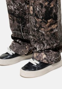 Camouflage-patterned loose-fit pants in earthy tones with tree and leaf designs, paired with black leather sneakers featuring white laces and a thick sole.