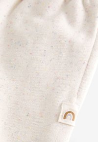 Tessuto di cotone morbido in beige chiaro con macchie multicolori. Presenta una piccola etichetta con un design arcobaleno dorato. Texture liscia.