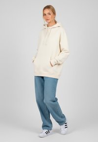 Felpa crema con cappuccio a cordino, tasca frontale e polsini a costine, abbinata a jeans larghi azzurri e sneakers bianche con strisce nere.