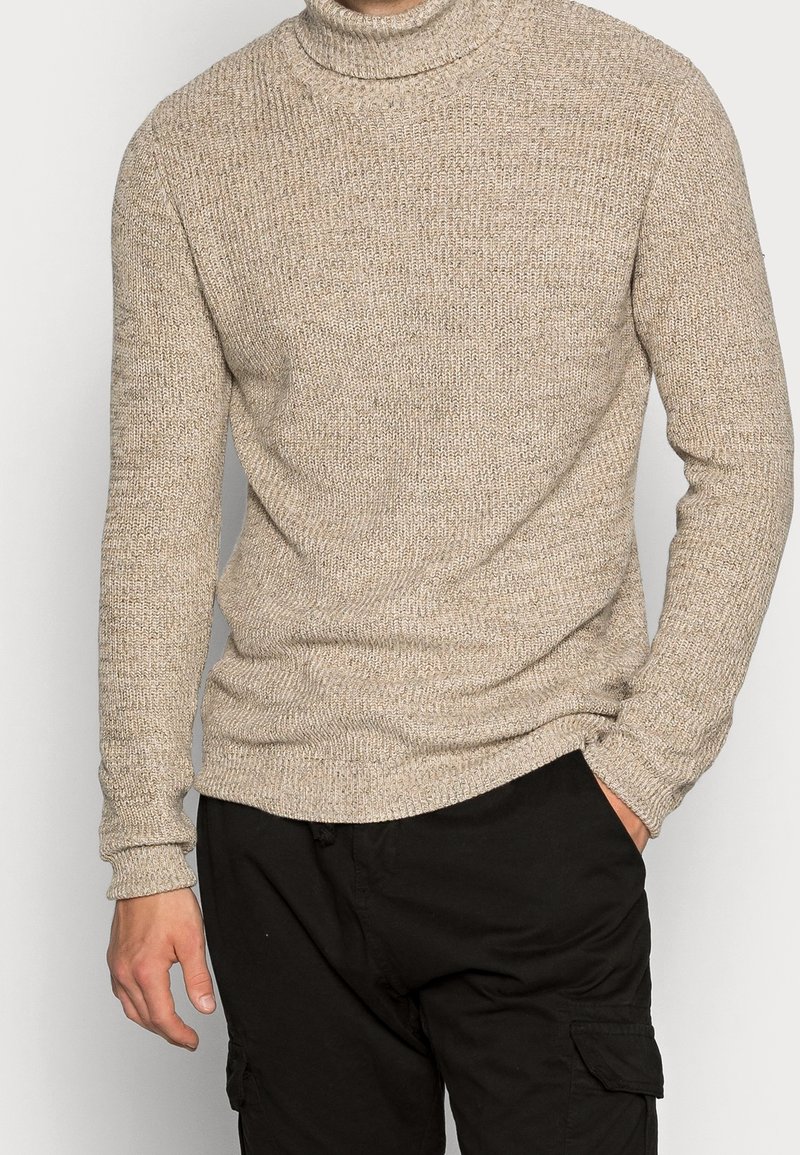 Beige gebreide coltrui met een gestructureerd oppervlak, aansluitende lange mouwen en een ribbenrand. Draag over donkere cargo-broeken.